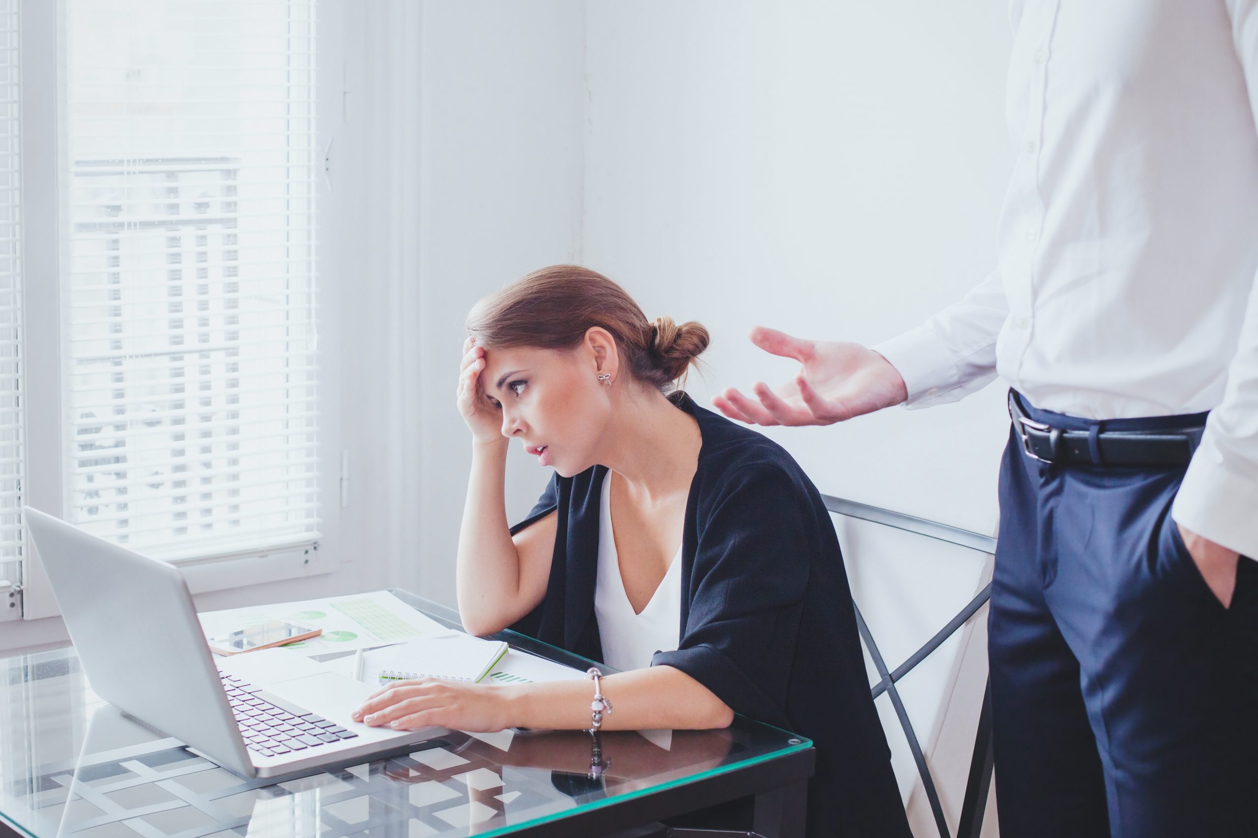 A woman is trying to work at her computer while her manager is adding more to her workload.