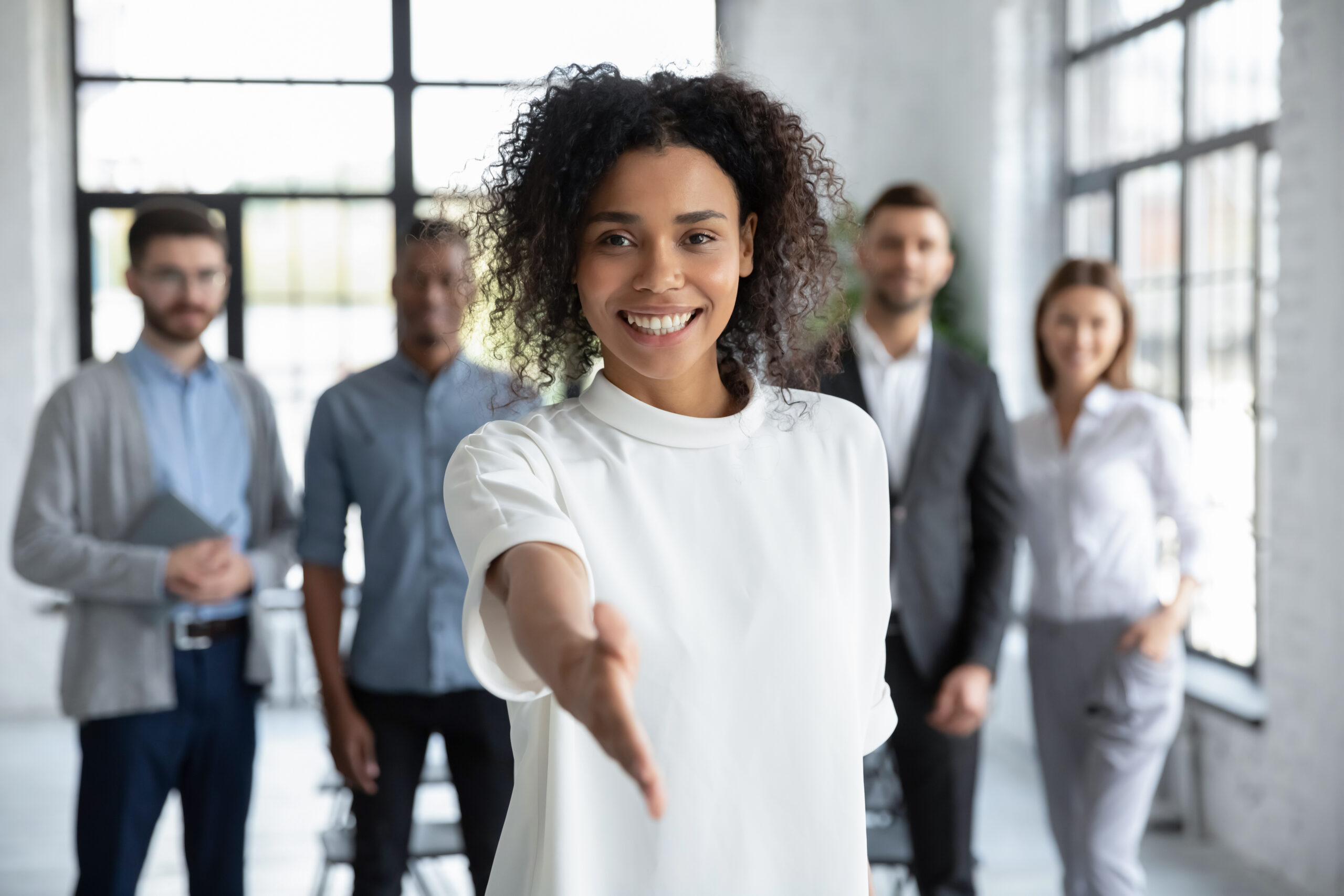 Black businesswoman standing in front of coworkers with her right hand extended, offering a handshake. [Have You Tried Being a Donor?]