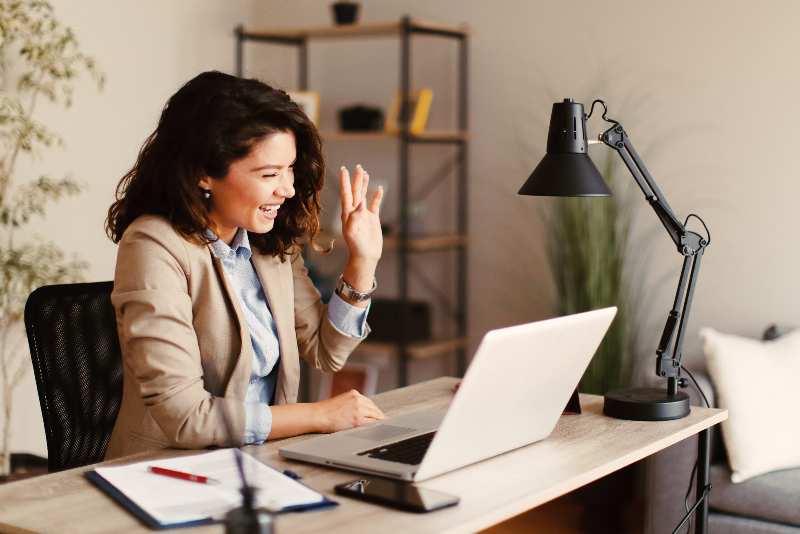 A young professional woman waves hello during a virtual meeting while sitting at her desk. [Stop Worrying About the Economy and Work Your Plan!]