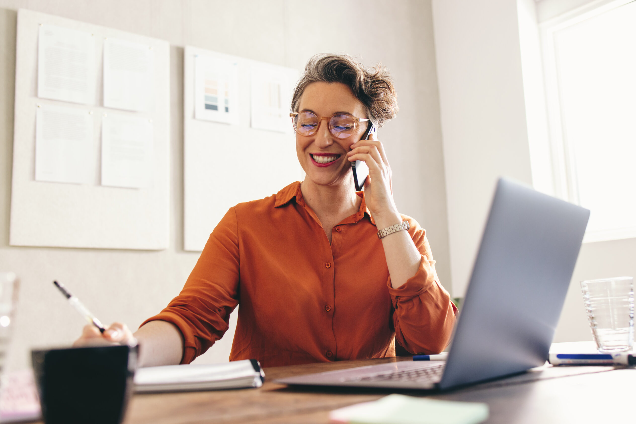 Smiling woman speaks on the phone while sitting at her desk with a notebook and laptop. [Two Simple Steps Helped a Donor Go from $50,000 to $1,000,000]