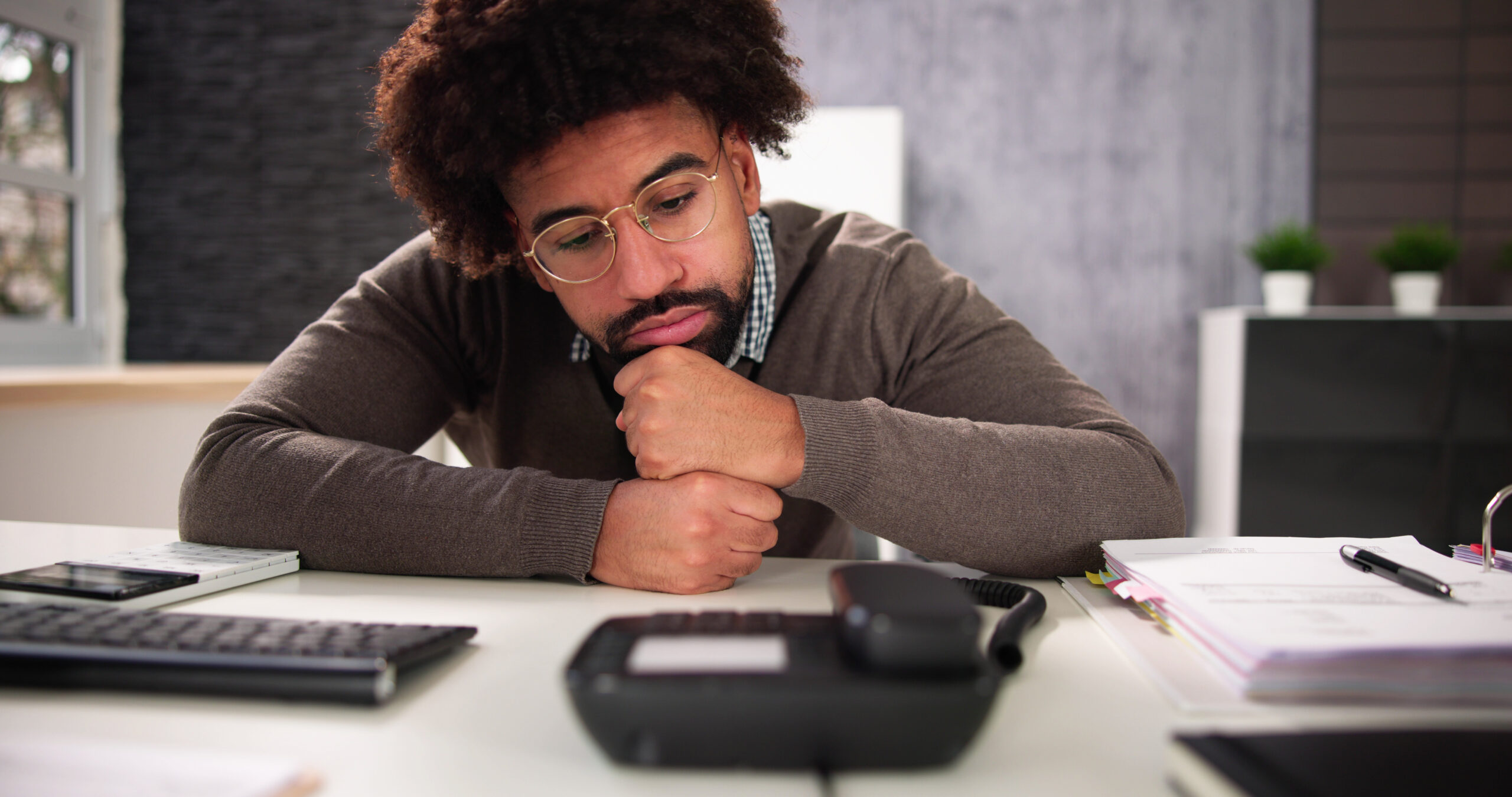 Professional man staring at landline phone on his desk while resting his chin on his hands. [The Stories We Tell about Donor Phone Calls]